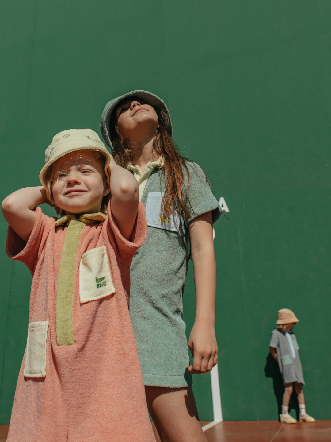 Peach | Three children wearing kids' jumpsuits: one in peach with lime green collar and placket, and yellow pockets; one in pale green with yellow collar and placket, with blue pockets; one in pale gray with blue collar and placket, and green pockets