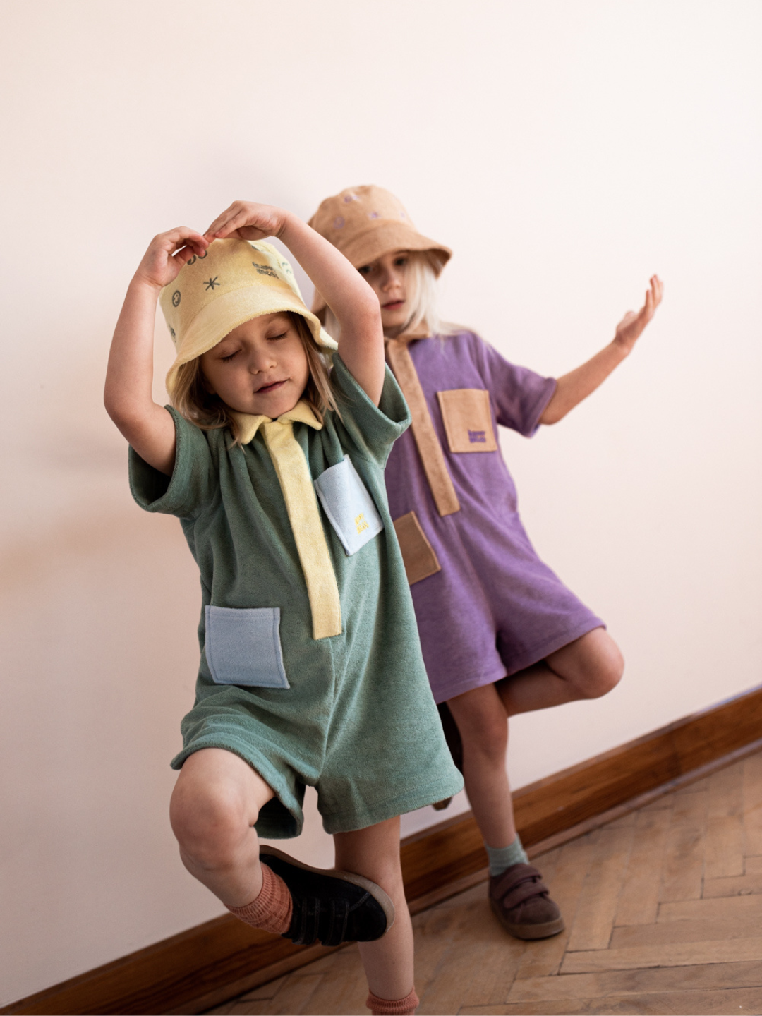 Plum | Two children dancing in kids' jumpsuits: one in pale green with yellow collar and placket and blue pockets; one in pale plum with sand collar, placket and pockets