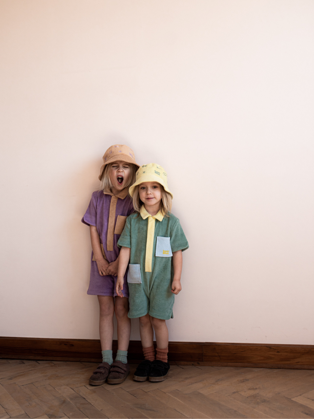Two children wearing kids' jumpsuits, one in pale plum with sand collar, placket and pockets; one in pale green with yellow collar and placket, and blue pockets