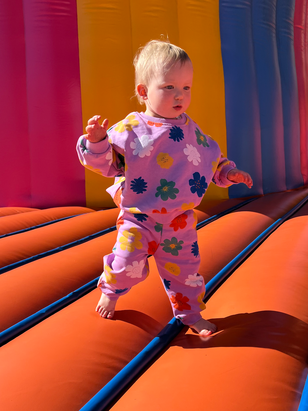 A toddler on a bouncy castle wearing a kids' sweatshirt and pants in purple, printed with yellow, orange, green, blue and white flowers
