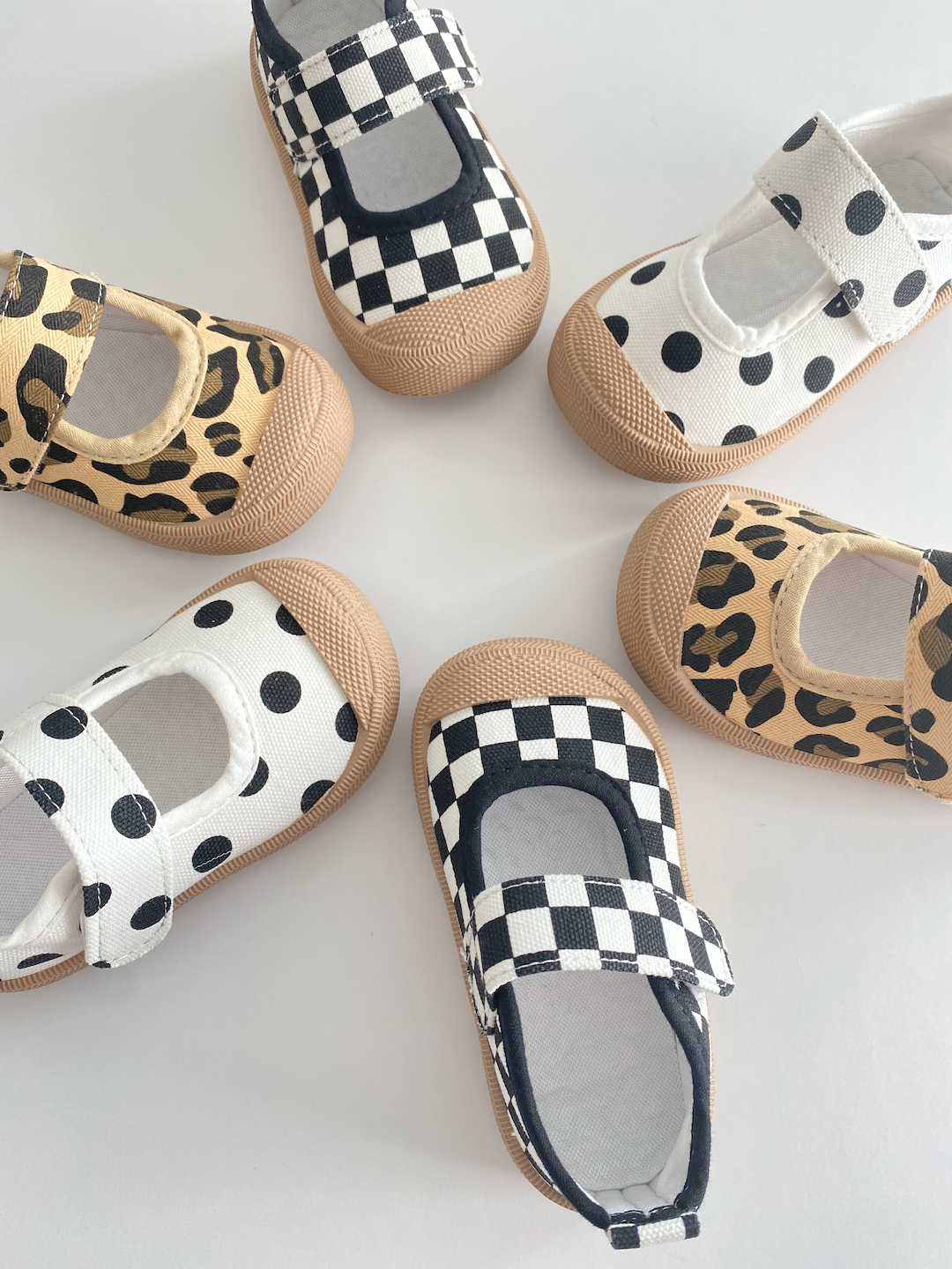 A selection of toddler mary jane sneakers arranged in a circle; various patterns: black-and-white checkerboard, white with black dots, leopard print