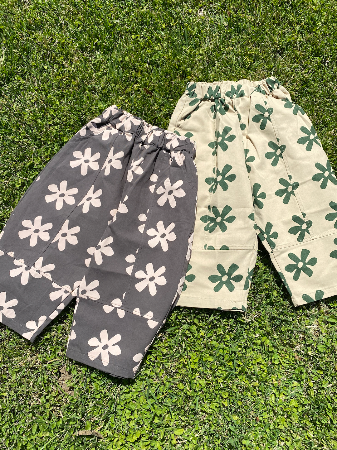 Two pairs of kids' pants laid on grass: one in gray with cream flowers, one in cream with green flowers