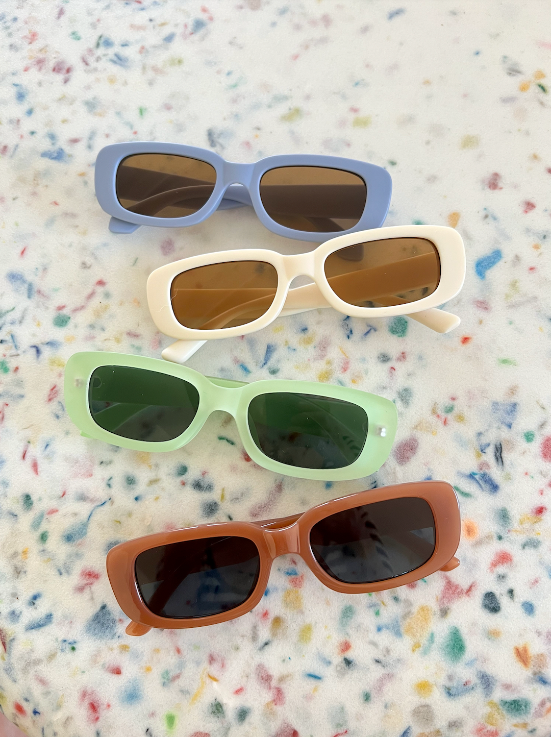Four pairs of kids' sunglasses: blue frames with brown lenses, cream frames with brown lenses, mint green frames with green lenses, orange frames with blue lenses