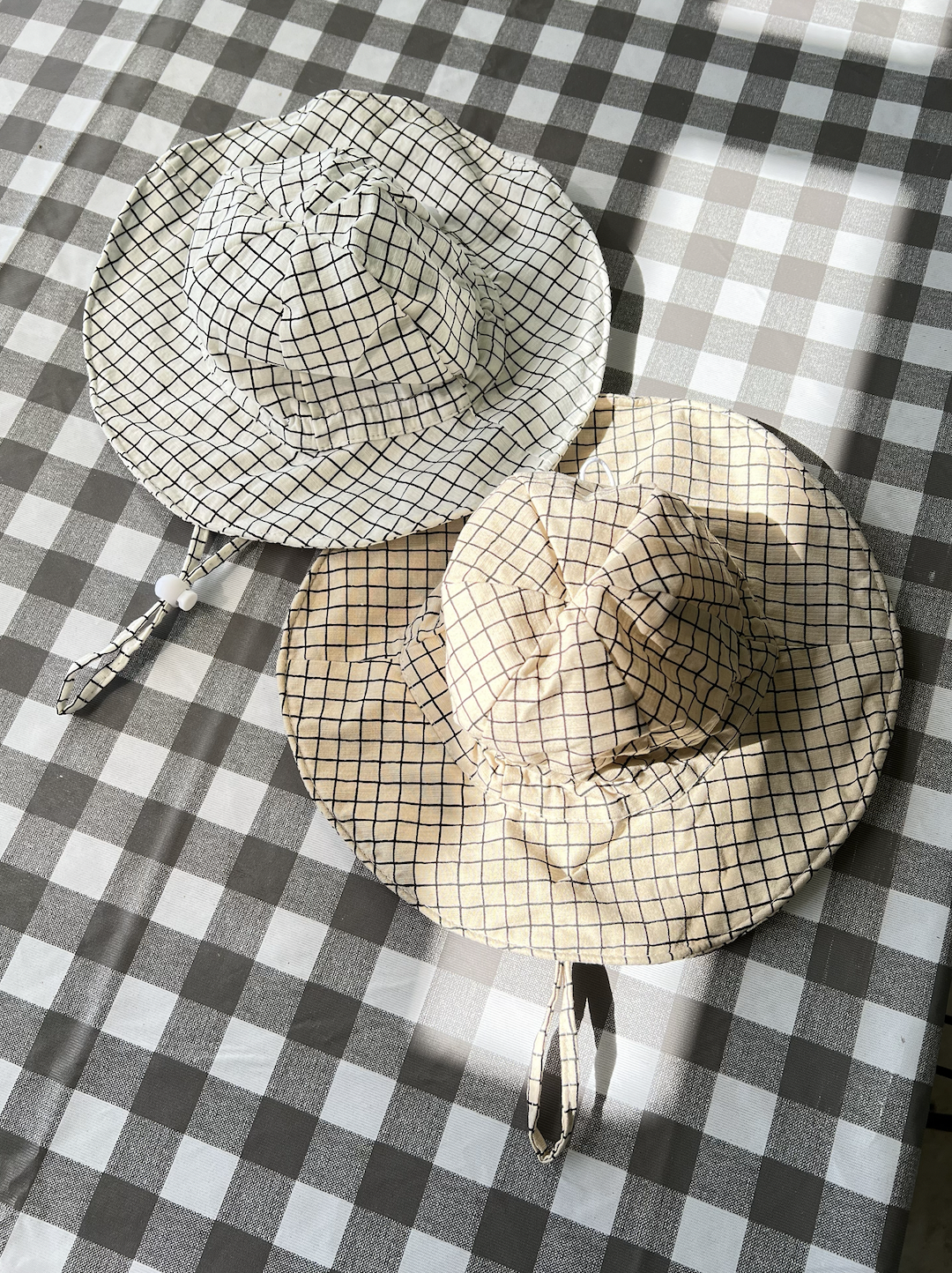 Two off the grid hats in latte and white are laid flat on a picnic check blanket