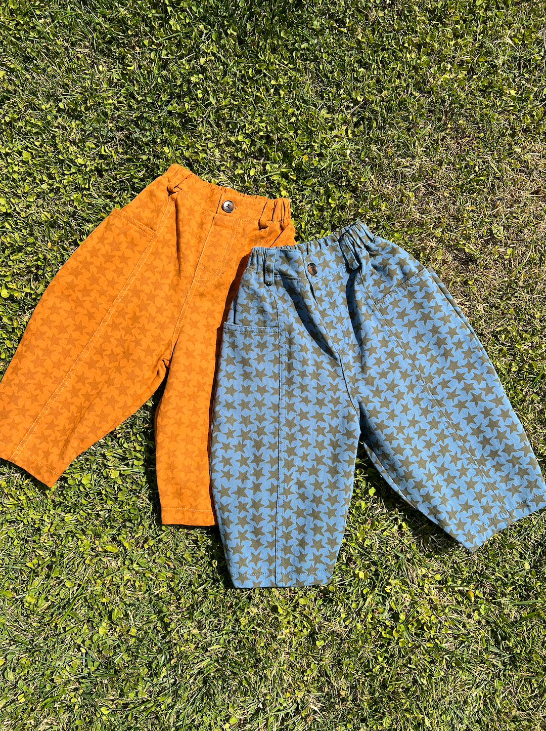 Two pairs of kids' pants laid on grass: one in orange with a rust star pattern, one in slate blue with olive green stars