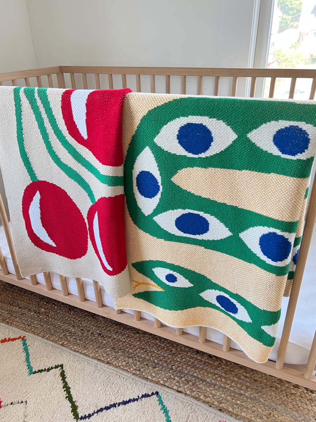 Two mini blankets folded over the edge of a cot, one in a red cherry pattern, one in a pattern of a green snake with blue eye markings