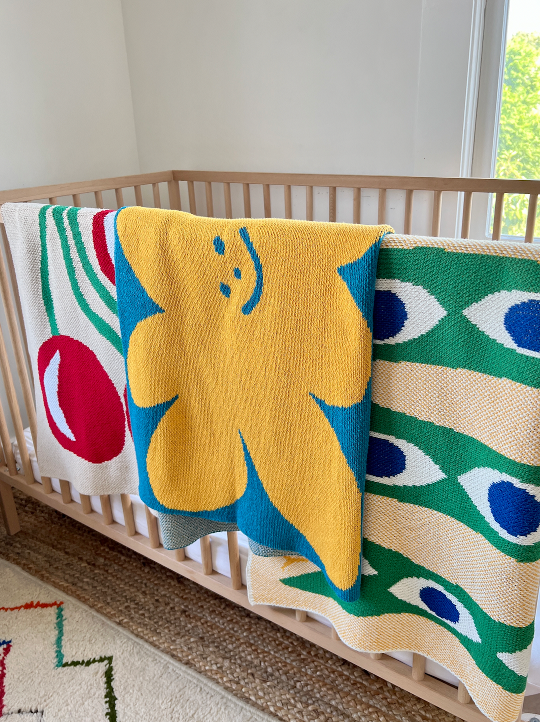 Three mini blankets folded over the side of a cot: one in a red cherry design, one with yellow and gray smiley shapes on a sky blue background, one with a green snake curling down an ecru background