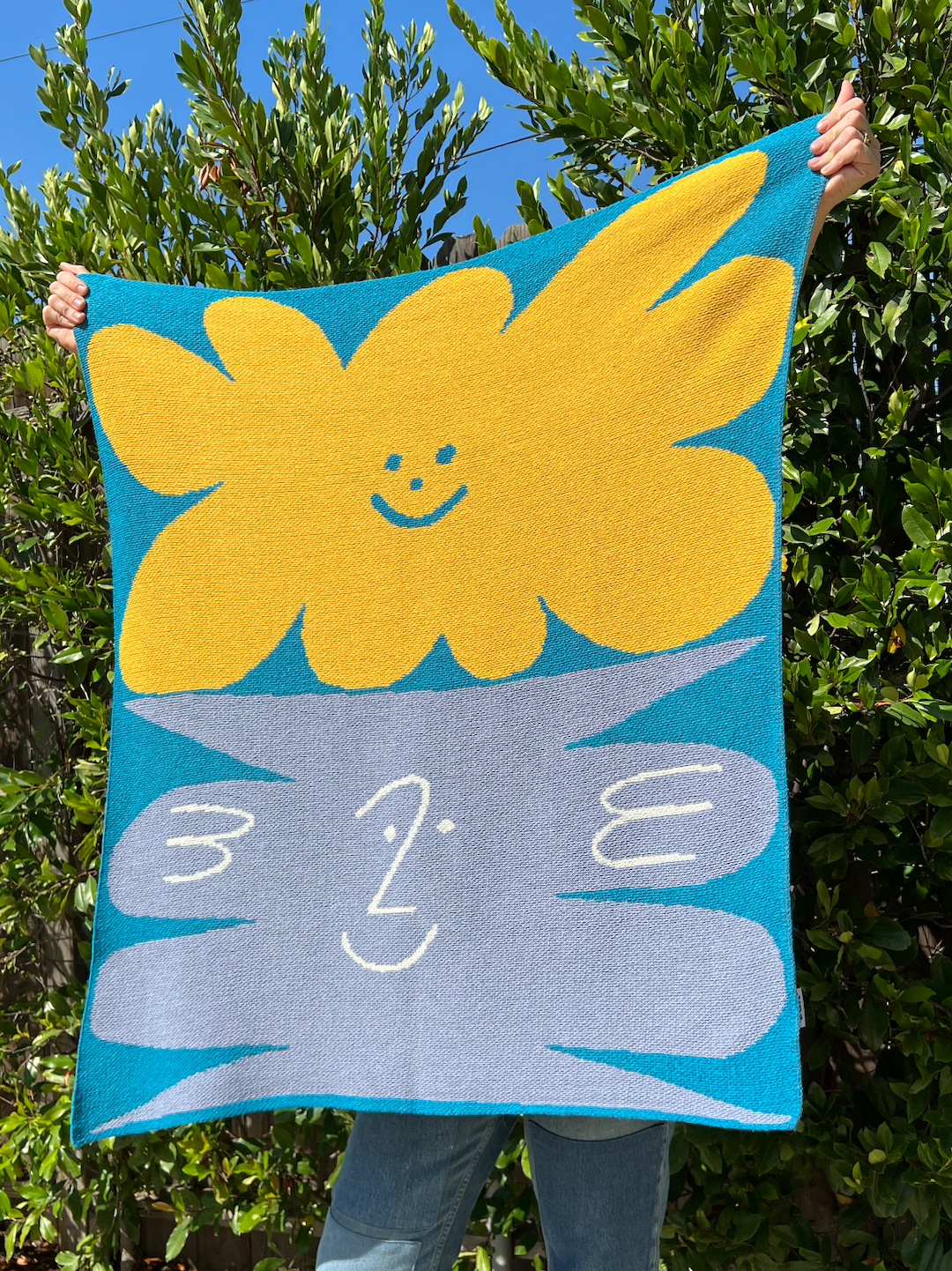 A mini blanket being held up for size, showing yellow and gray smiley patterns on a sky blue background
