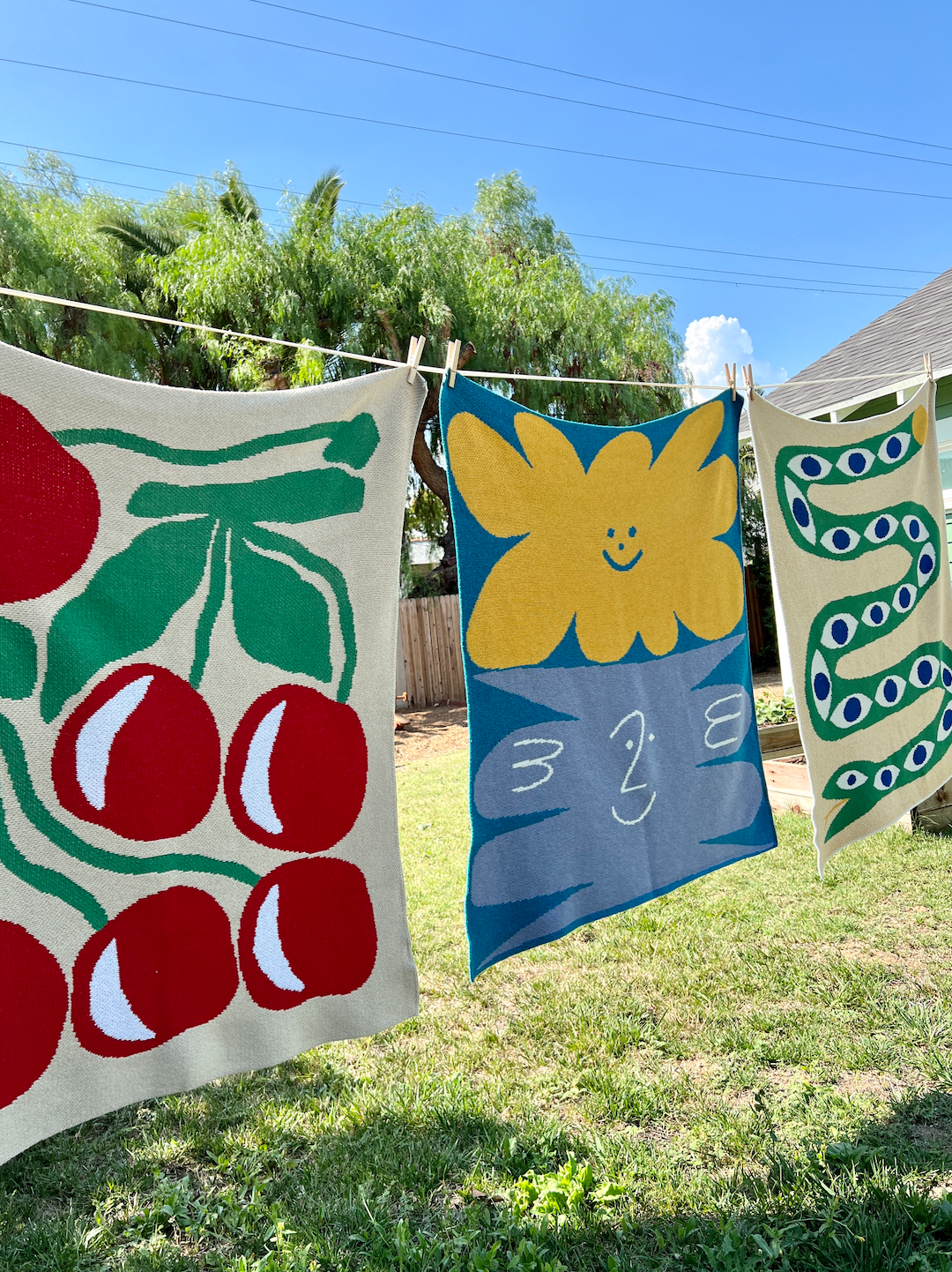 Three mini blankets on a washing line: one with red cherries and green leaves on an ecru background, one with yellow and gray smiley shapes on a sky blue background, one with a green snake with blue eye patterns curling down an ecru background