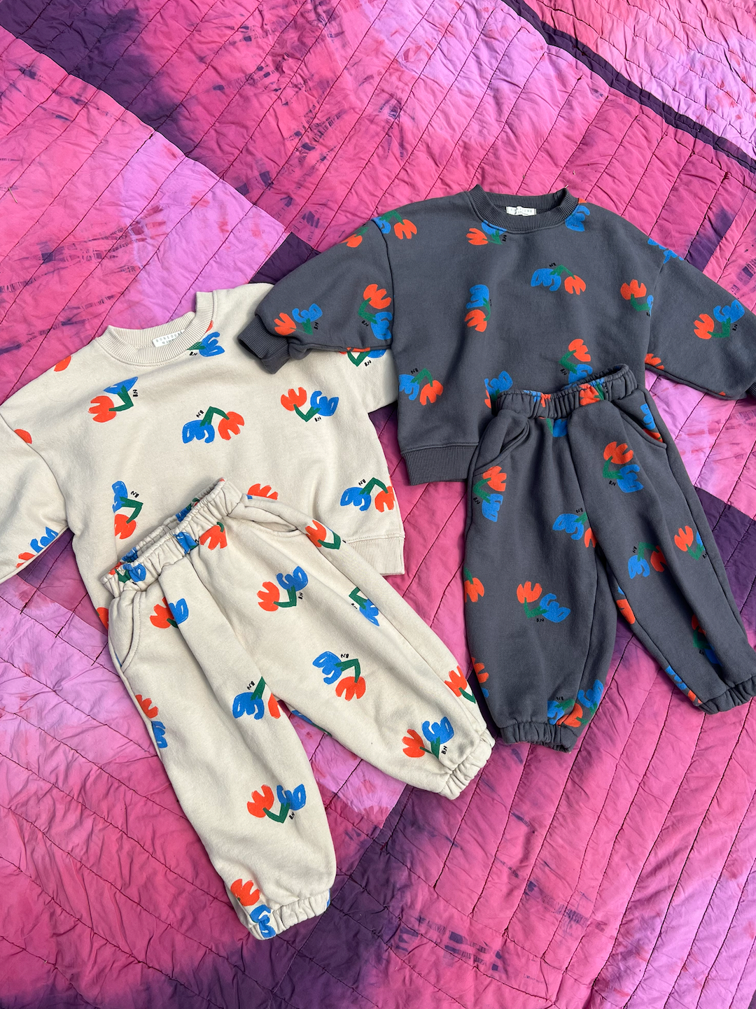 Two pairs of kids' sweatpants and tops laid on a quilt. One pair with blue and red flowers on an ecru background, one pair with blue and orange flowers on a slate gray background