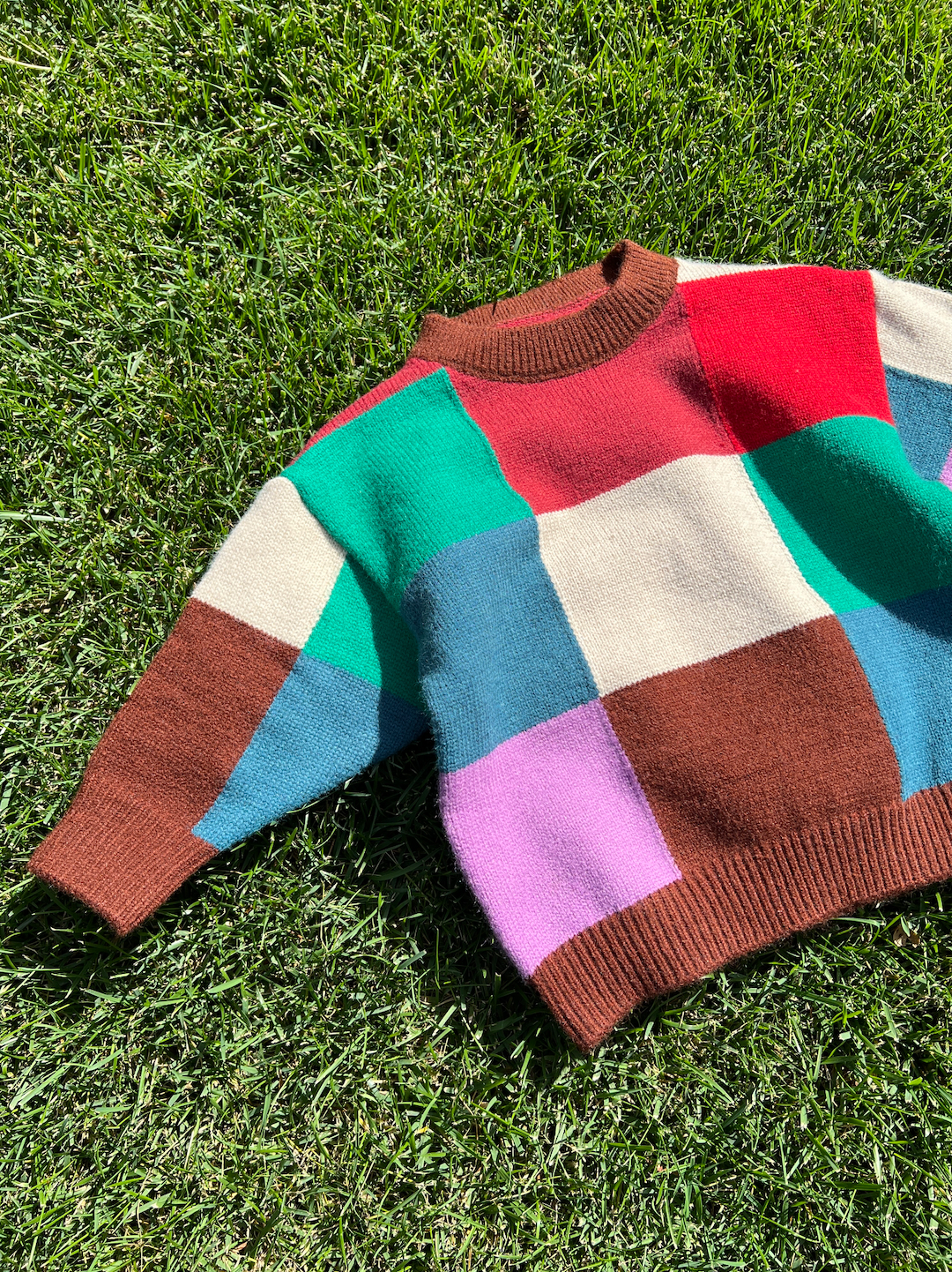 A kids' colorblock sweater laid on grass, in shades of red, pink, cream, brown, blue and green