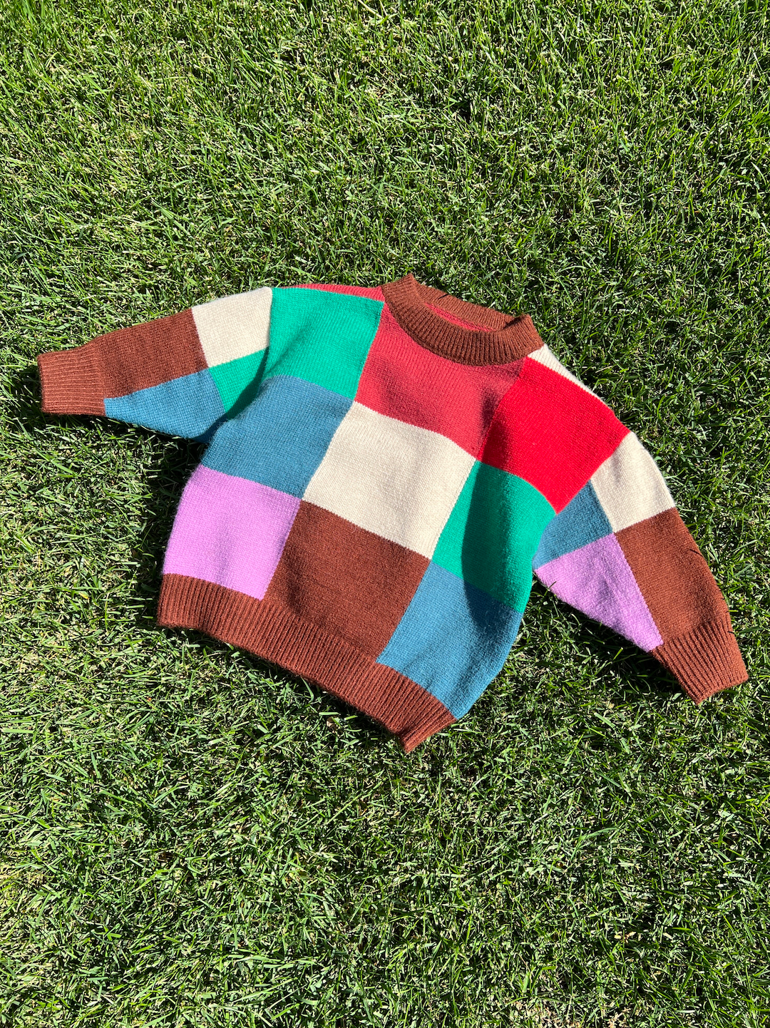 A kids' colorblock sweater laid on grass, in shades of red, pink, cream, brown, blue and green