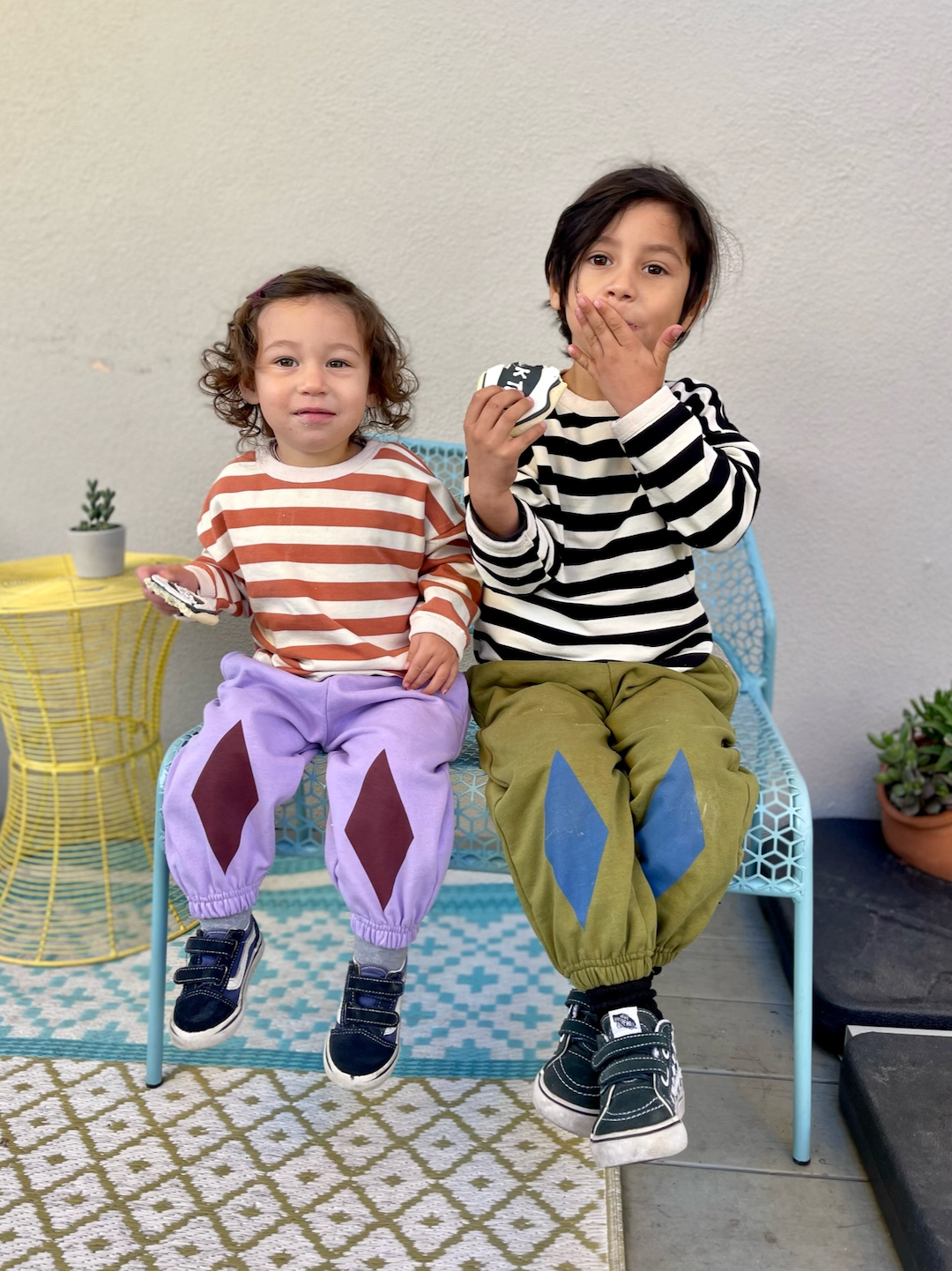 Two children sitting on a bench in kids' sweatpants; one pair in pale purple with dark red diamond patches on the knees, one in olive green with blue diamond patches