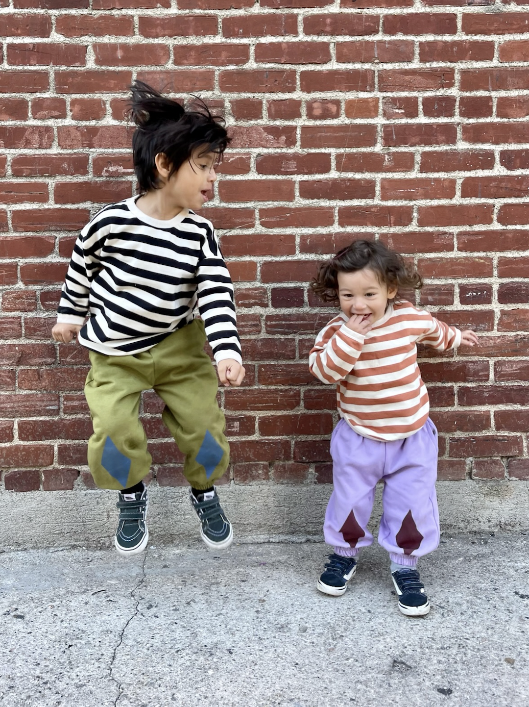 Two children jumping in kids' sweatpants; one in olive green with blue diamond patches at the knees; one in pale purple with dark red diamonds