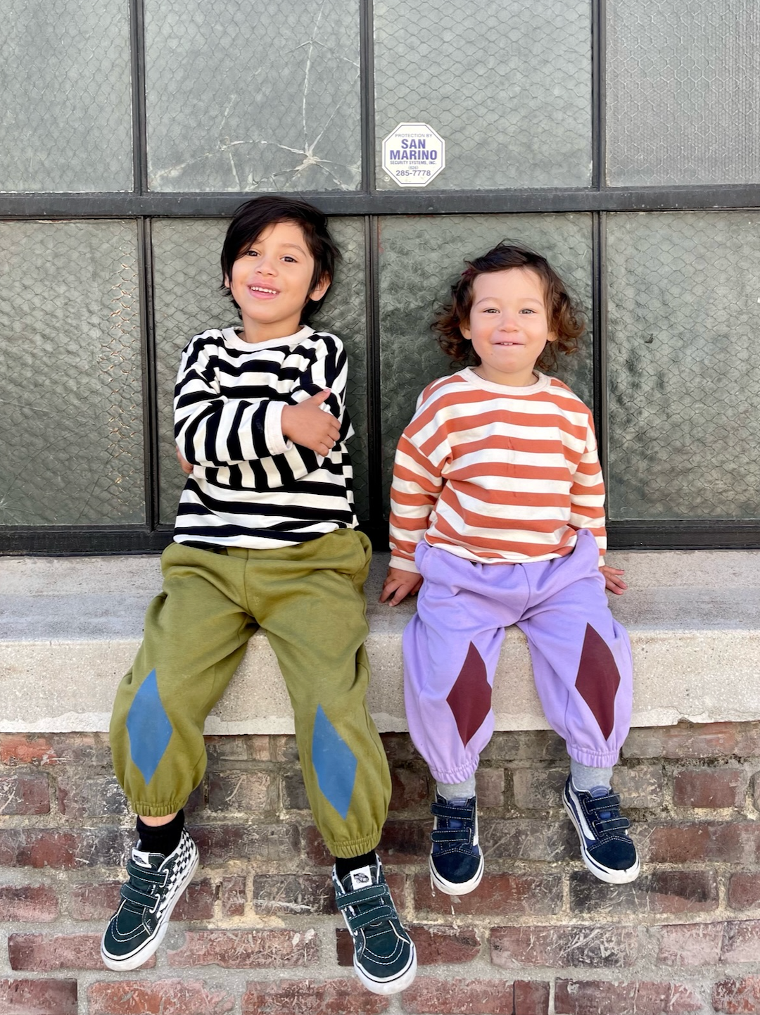 Two children sitting on a wall wearing kids' sweatpants; one in olive green with blue diamond patches at the knees, one in pale purple with dark red diamonds
