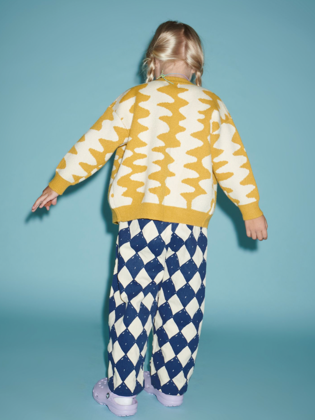 Marigold | A girl standing facing backwards, wearing Marigold Wavelength Cardigan with diamond pants and purple crocs. 