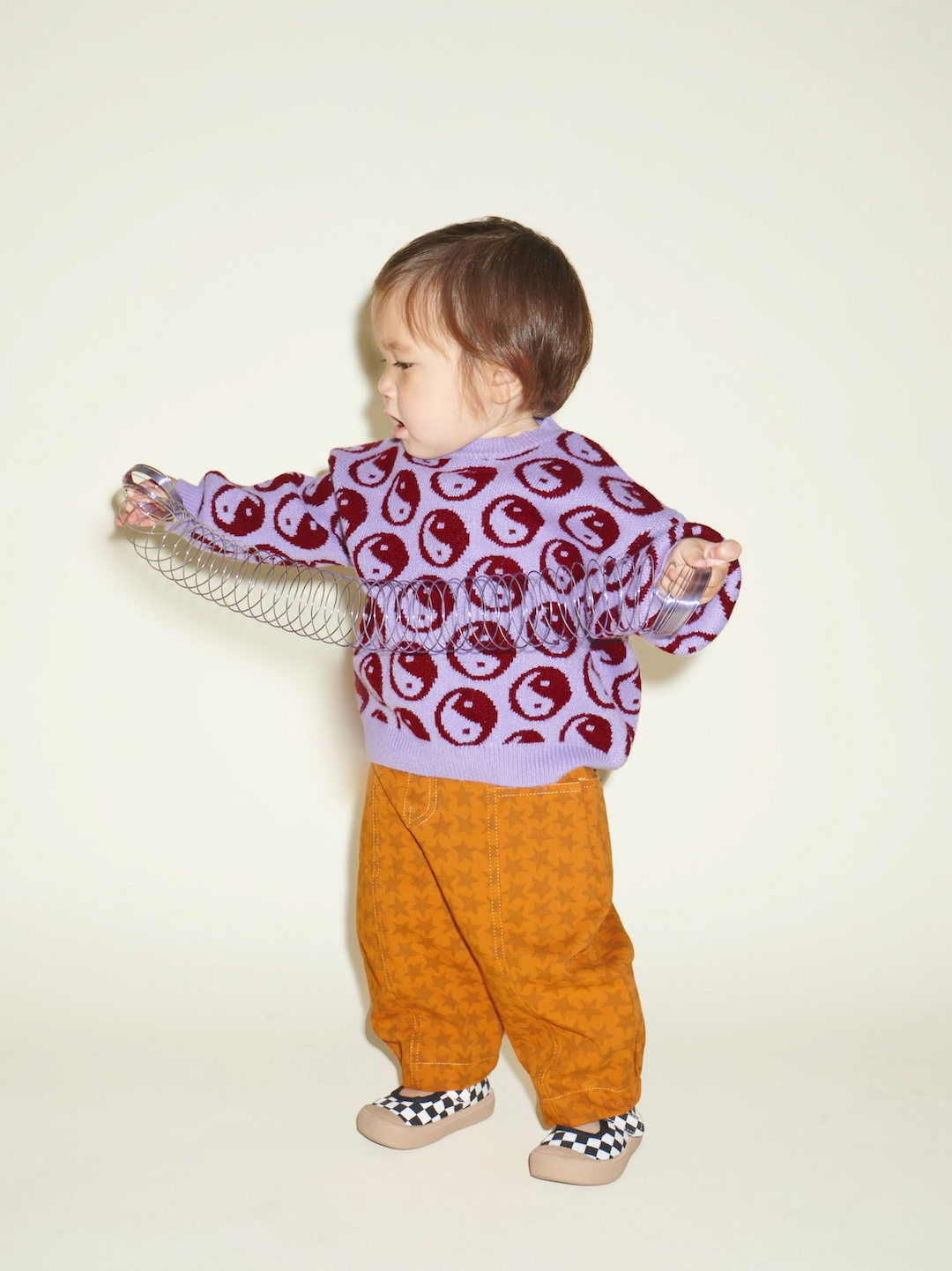 A toddler with a slinky wearing a pair of kids' pants in an orange and rust star pattern, under a Cosmos Sweater in violet