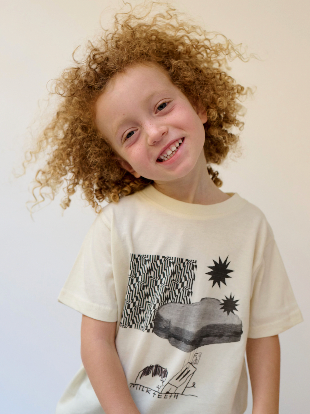 A smiling child wearing a kids' tee shirt in cream, printed in black with stars, zigzags, a PBJ sandwich, a toaster and the words Milk Teeth
