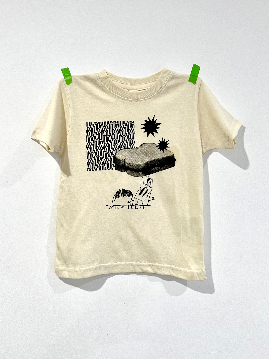 A kids' tee shirt in cream, printed in black with zigzag patterns, stars, a PBJ sandwich, a toaster and the words Milk teeth