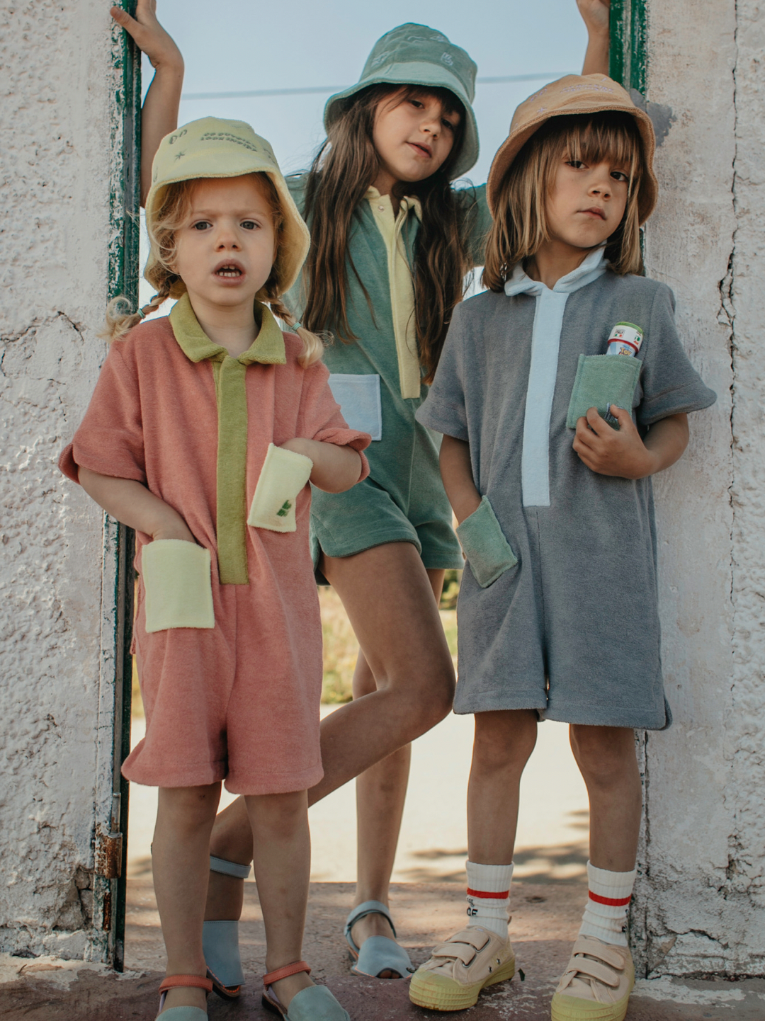Stone | Three children wearing kids' jumpsuits in different colors: one peach with green collar and placket, one green with yellow collar and placket, one pale gray with blue collar and placket