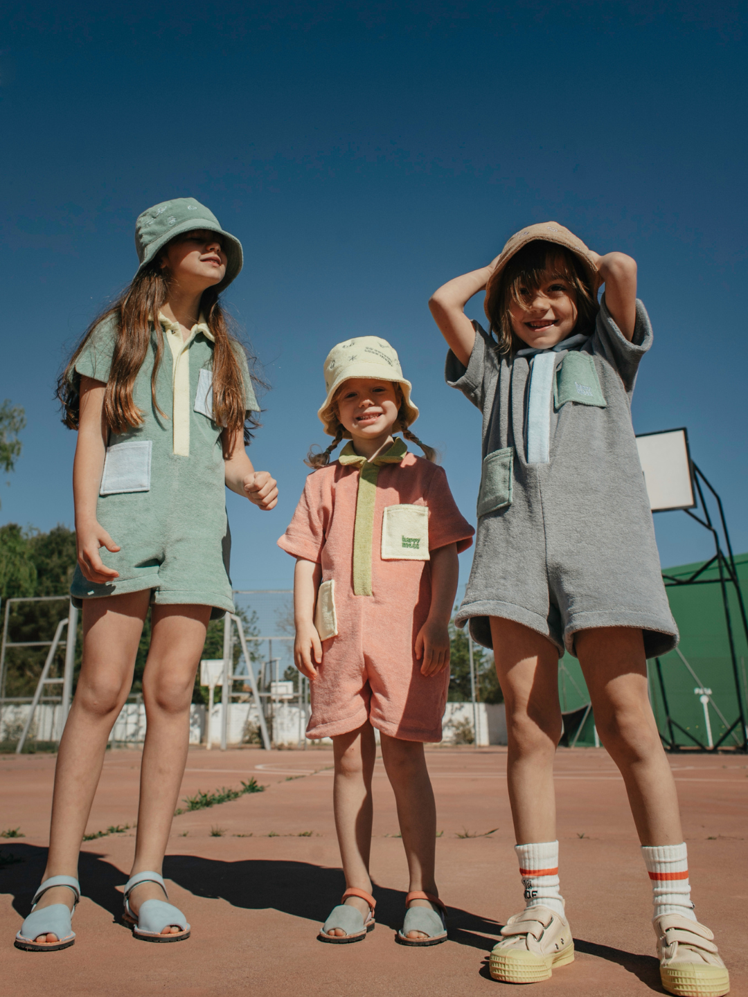 Stone | Three children wearing kids' jumpsuits in different colors: one pale green with yellow collar and placket, and blue pockets; one in peach with lime green collar and placket, and yellow pockets; one in pale gray with blue collar and placket, and green pockets