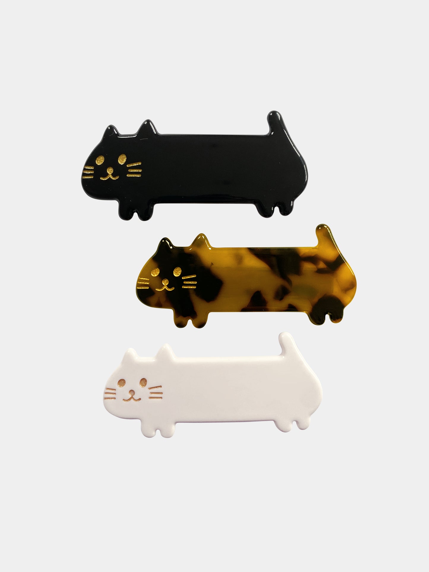 A set of three kids' barrettes in the shape of elongated cats; one black, one tortoiseshell, one white